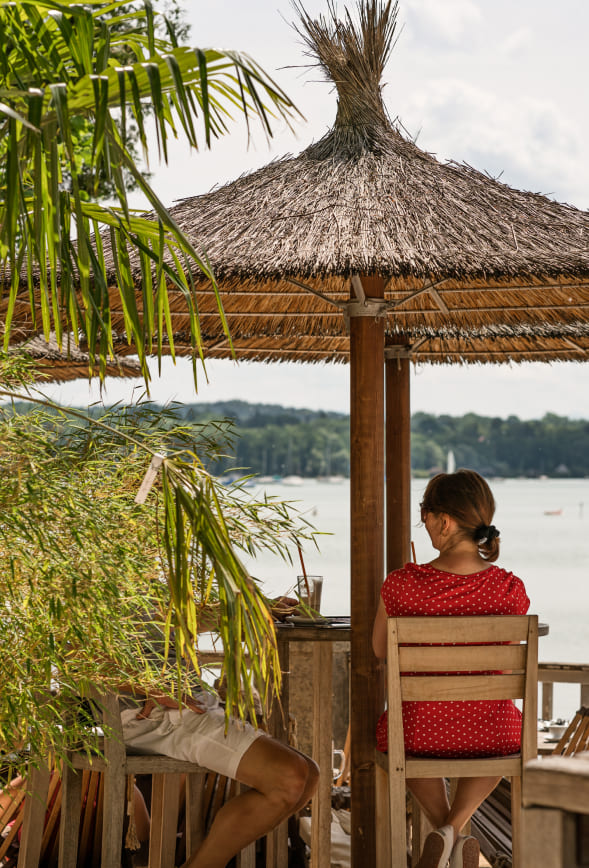 Gäste sitzen unter Strohschirm der Strandbar mit Blick auf den Ammersee